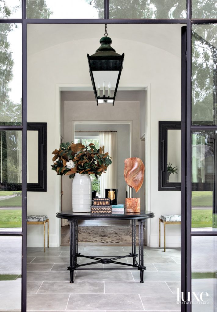 Custom steel entry doors from Atelier Domingue lead to a foyer where a 19th-century French lantern from W. Gardner Antiques lights an antique table from Shabby Slips Home and sculpture from Found. Mirrors—also from W. Gardner Antiques—overlook benches in Tillett Textiles linen. The limestone flooring is from Farmhouse Stone.